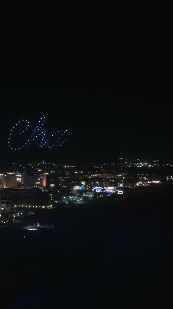 Letra cursiva dibujada por un enjambre de drones en el cielo nocturno sobre las luces de un hotel frente a la bahía